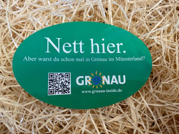 Ovaler, grüner Aufkleber mit der Aufschrift "Nett hier, aber warst du schon mal in Gronaui im Münsterland?", einen QR-Code und dem Logo des Stadtmarketings Gronau. Im Hintergrund ist Stroh zu sehen.