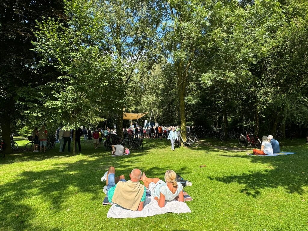 Twee mensen liggen op een deken in de schaduw en kijken naar een groepje mensen onder de bomen in een zonnig park. Vlakbij staan fietsen geparkeerd; anderen zitten of staan in het gras en genieten van het evenement in de open lucht.
