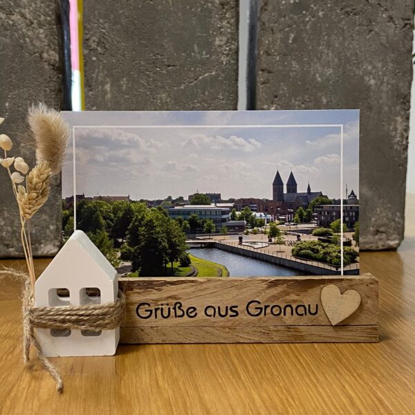 Ein Kartenständer mit der Aufschrift "Grüße aus Gronau" mit einem Haus aus Gips und Trockenblumen als Dekoration, in dem eine Postkarte aus Gronau steht.