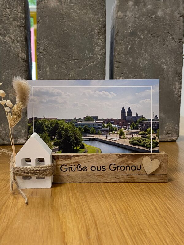 Ein Kartenständer mit der Aufschrift "Grüße aus Gronau" mit einem Haus aus Gips und Trockenblumen als Dekoration, in dem eine Postkarte aus Gronau steht.