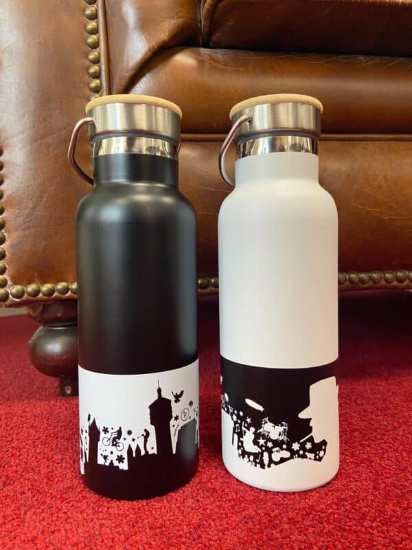 Eine schwarze Gronau Trinklasche mit weißem Motiv und eine weiße Gronau-Trinkflasche mit schwarzem Motiv nebeneinander auf einem roten Untergrund.
