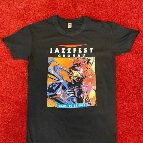 Das Bild zeigt ein schwarzes T-Shirt, das flach auf einem roten Teppich liegt. Auf der Vorderseite des Shirts steht oben groß „JAZZFEST GRONAU“. Darunter befindet sich ein buntes, abstraktes Kunstmotiv in kräftigen Farben wie Orange, Blau, Pink und Gelb. Am unteren Rand des Motivs steht ein blaues Datumsfeld mit dem Zeitraum „28.04.–03.05.2026“.