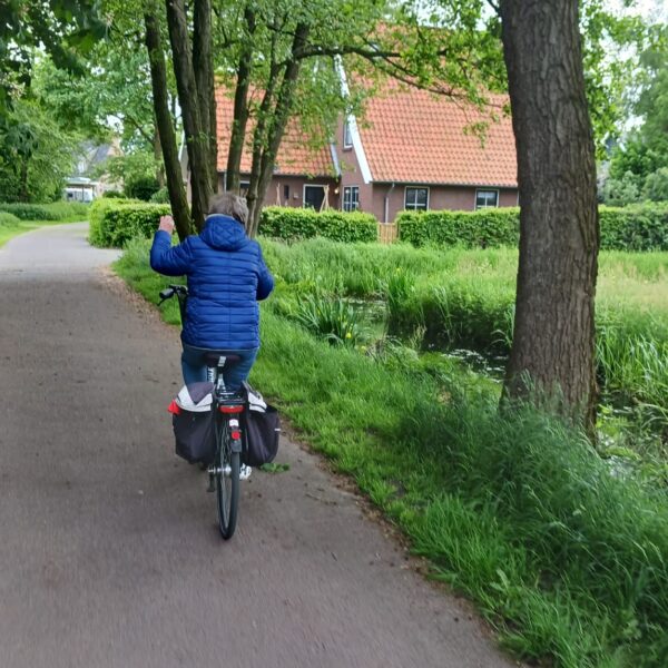 Das Foto zeigt eine Person, die auf einem Fahrrad auf einem asphaltierten Weg fährt. Die Person trägt eine blaue Steppjacke und hat graue Haare. Links und rechts des Weges stehen viele grüne Bäume und Büsche. Rechts vom Weg befindet sich ein kleiner Graben mit Wasser und dichtem Gras. Im Hintergrund ist ein Haus mit rotem Ziegeldach zu sehen, teilweise verdeckt von den Bäumen.