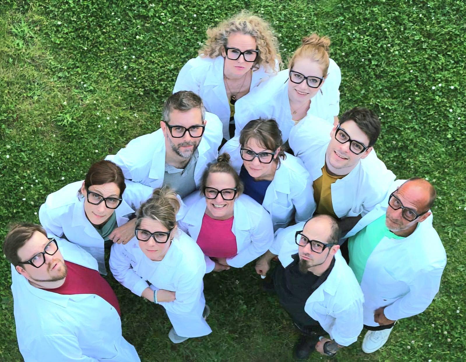 Eine von oben fotografierte Gruppe von 11 Personen, die in die Kamera lächeln. Allesamt tragen sie schwarze Brillen, bunte Shirts und weiße Kttel.