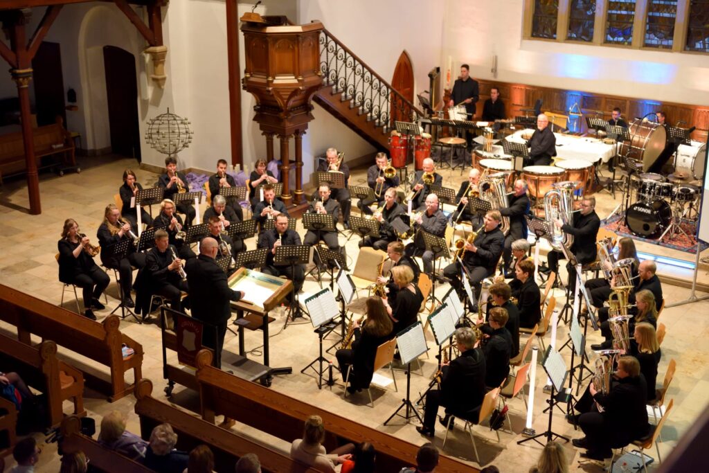 Ein großes Orchester spielt in einer Kirche, wobei die Musiker verschiedene Blas- und Schlaginstrumente spielen. Der Dirigent steht vorne, und das Publikum sitzt in den hölzernen Kirchenbänken und beobachtet die Aufführung.