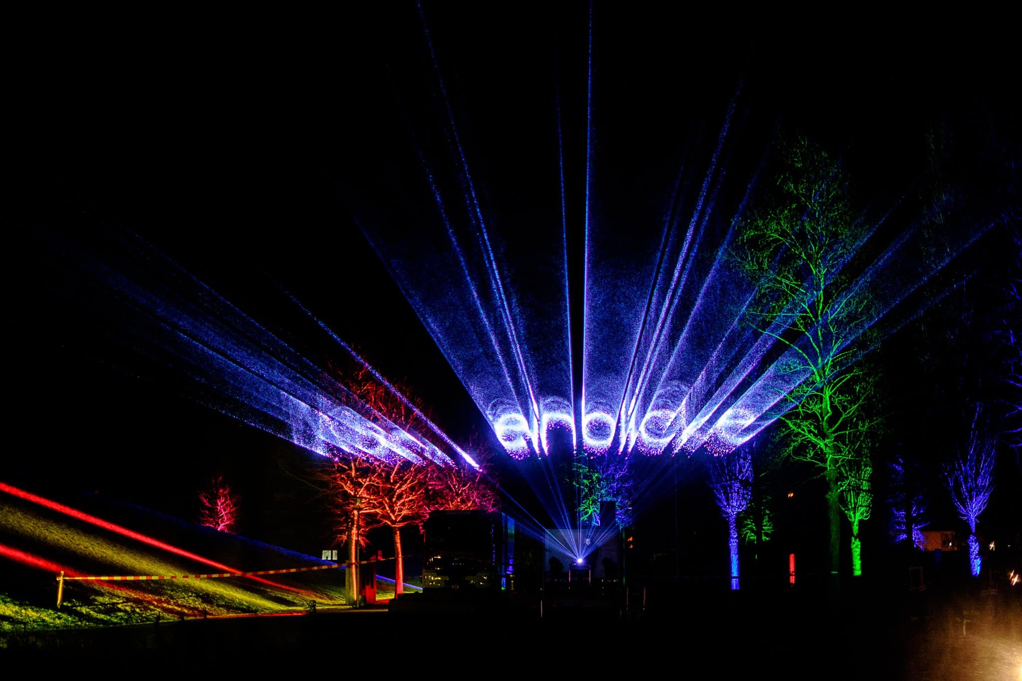 Colorful lasers project the words "Night Insights" into the dark night sky and illuminate the surrounding trees in red, blue and green hues during an outdoor light show.