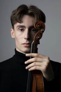 Ein junger Mann mit braunen Haaren und schwarzem Rollkragenpullover hält eine Geige aufrecht neben sein Gesicht, wirbt für das Kammerkonzert mit dem Duo Darius Preuß und János Palojtay und blickt in die Kamera vor einem schlichten grauen Hintergrund.