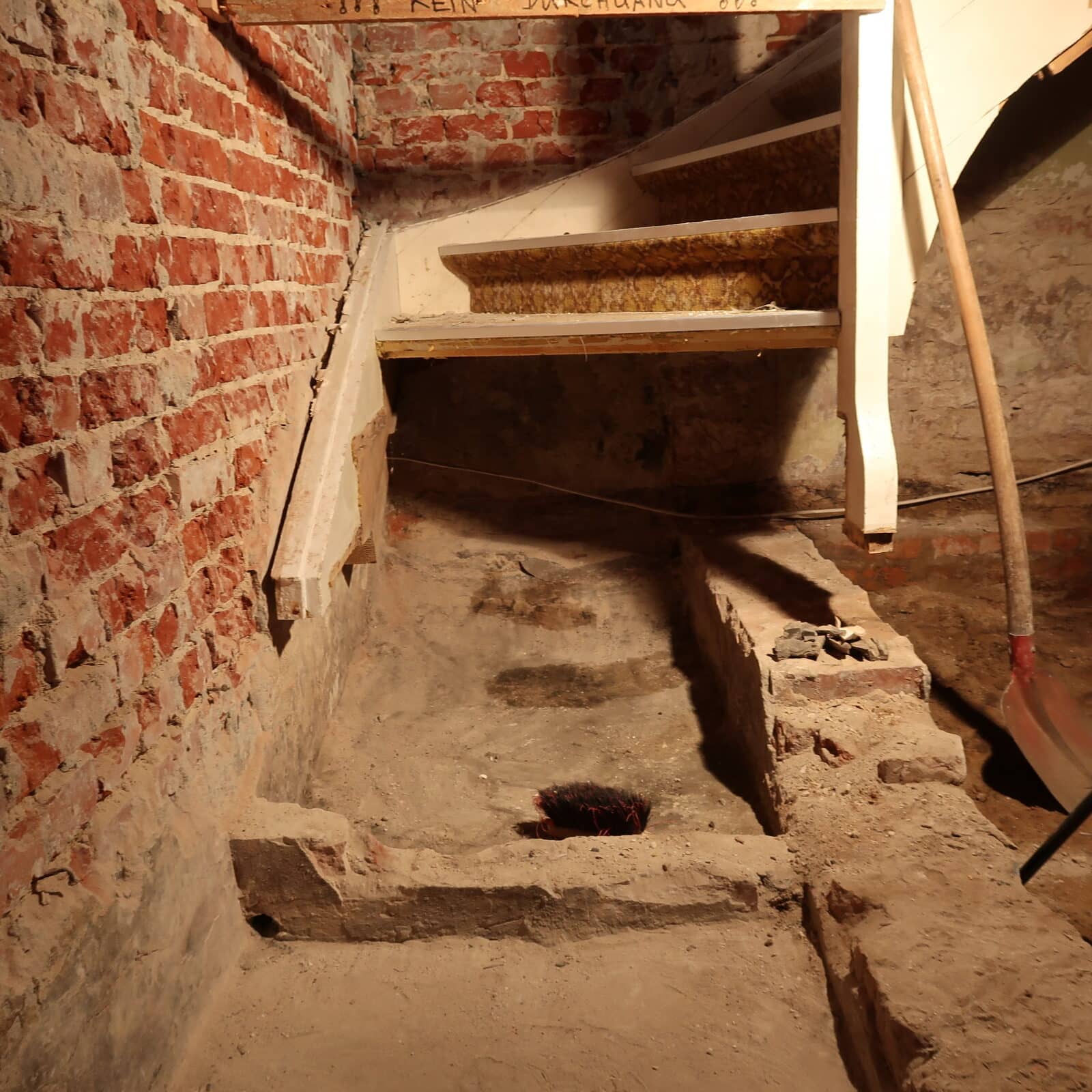 Das Foto zeigt einen alten, staubigen Kellerbereich aus rotem Ziegelmauerwerk. Eine steile, helle Holztreppe führt nach oben. Im Vordergrund ist der Boden teilweise aufgebrochen, mit freigelegtem Mauerwerk und Erde. Ein Besen liegt am Boden, daneben steht eine Schaufel an der Wand. Die Beleuchtung ist warm und schwach, wodurch der Raum alt und etwas verlassen wirkt.