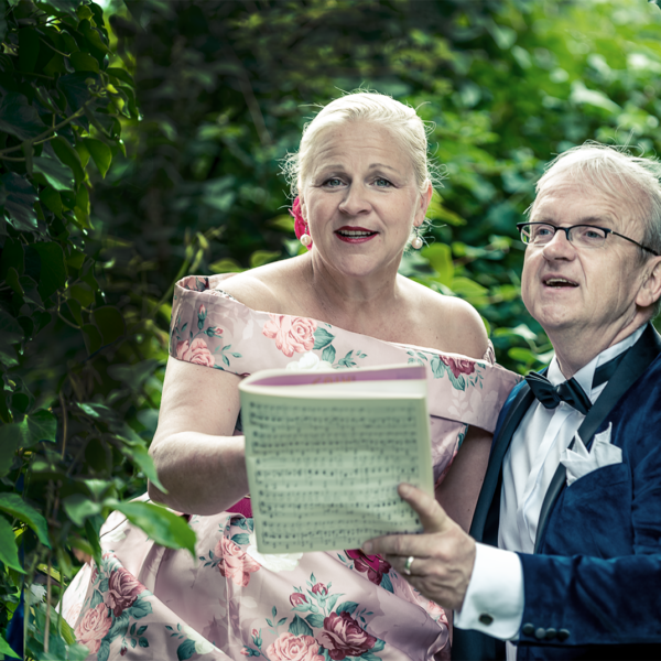 Zwei Personen stehen nah beieinander inmitten dichter grüner Blätter, als wären sie in einem Garten. Links steht eine Frau im festlichen, rosafarbenen Kleid mit Blumenmuster. Rechts steht ein Mann im dunkelblauen Samtjackett und Fliege. Beide halten gemeinsam ein aufgeschlagenes Notenheft und blicken lächelnd nach vorn, als würden sie singen oder etwas präsentieren. Die Umgebung wirkt ruhig und natürlich.