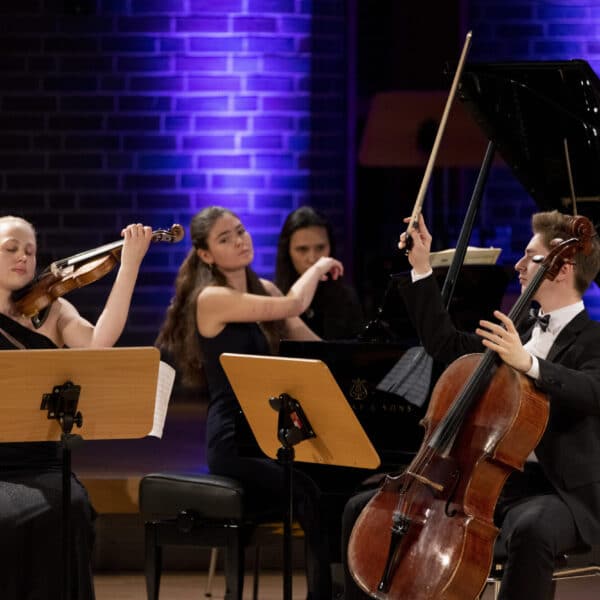 Auf dem Bild spielt ein Kammermusik-Trio auf einer Bühne. Links sitzt eine Geigerin, die mit geschlossenen Augen und erhobenem Bogen sehr konzentriert wirkt. In der Mitte sitzt eine Pianistin am Flügel, ebenfalls vertieft ins Spiel. Rechts spielt ein Cellist im Anzug, der seinen Bogen ebenfalls hochhebt. Hinter ihnen leuchten violette Scheinwerfer vor einer Backsteinwand. Die Stimmung wirkt feierlich und intensiv.