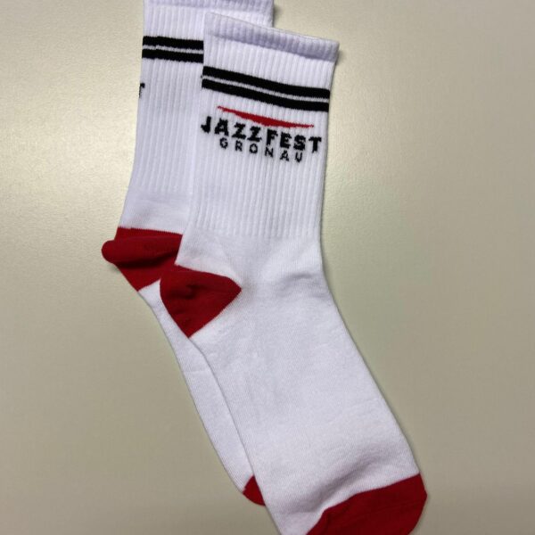 Auf dem Foto liegen zwei weiße Socken übereinander auf einer glatten, beigefarbenen Oberfläche. Die Socken haben rote Zehen- und Fersenbereiche sowie einen roten Streifen am oberen Fußbereich. Am Schaft befinden sich zwei dünne schwarze Streifen. Zwischen den Streifen ist das Logo „Jazzfest Gronau“ eingestickt – das Wort „Jazzfest“ in schwarzen Buchstaben mit einem roten, leicht geschwungenen Strich darüber, darunter kleiner der Schriftzug „Gronau“. Die Socken wirken neu und ordentlich zusammengelegt.