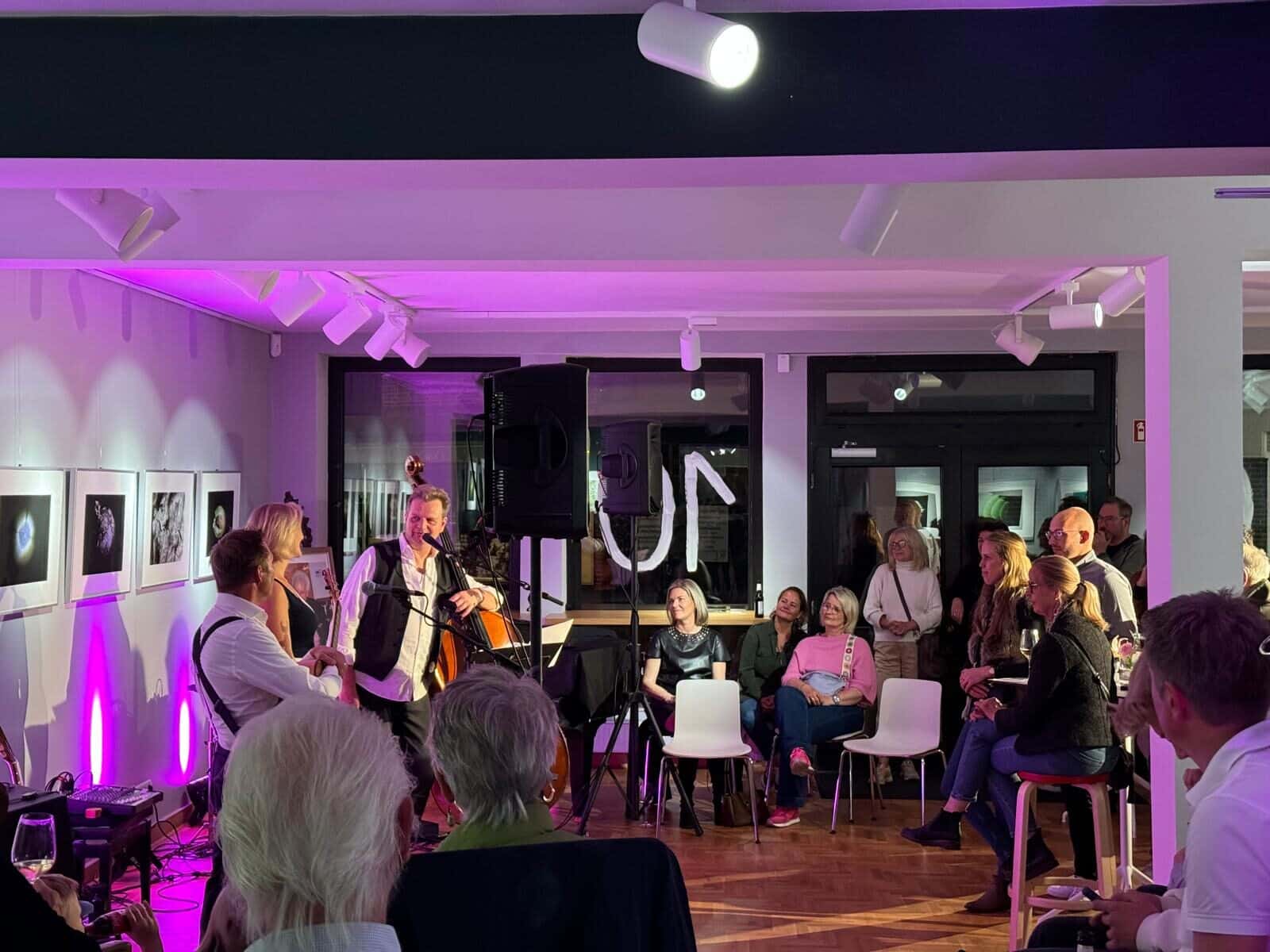 In einem hellen Raum findet ein kleines Live-Konzert statt. Drei Musiker stehen im Mittelpunkt, einer davon spielt ein großes Streichinstrument (Kontrabass). Um sie herum sitzen und stehen zahlreiche Zuhörerinnen und Zuhörer auf Stühlen und an den Wänden. Die Atmosphäre ist gemütlich, mit violett-rosa Beleuchtung, Bildern an der Wand und Scheinwerfern an der Decke. Das Publikum schaut aufmerksam zu.