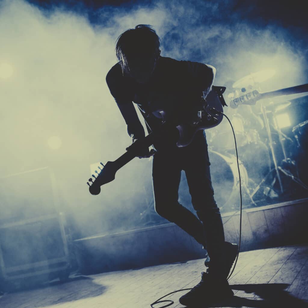 Foto van een gitarist. Je ziet alleen schaduwen op een podium. Donkerblauwe achtergrond met mist.