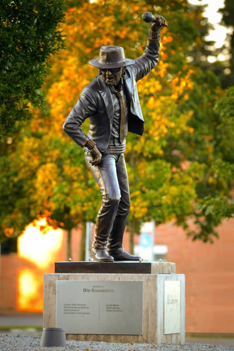 Bronzen standbeeld van een zanger op een stenen sokkel in een park. Het figuur draagt een hoed, een zonnebril, een jasje en een strakke broek. In haar rechterhand houdt ze een microfoon omhoog, alsof ze net op het podium staat op te treden. De pose oogt dynamisch en energiek. Op de achtergrond zijn vage bomen te zien in warme herfstkleuren: geel, oranje en groen. Op de sokkel staat het opschrift „Die Gronauten“.