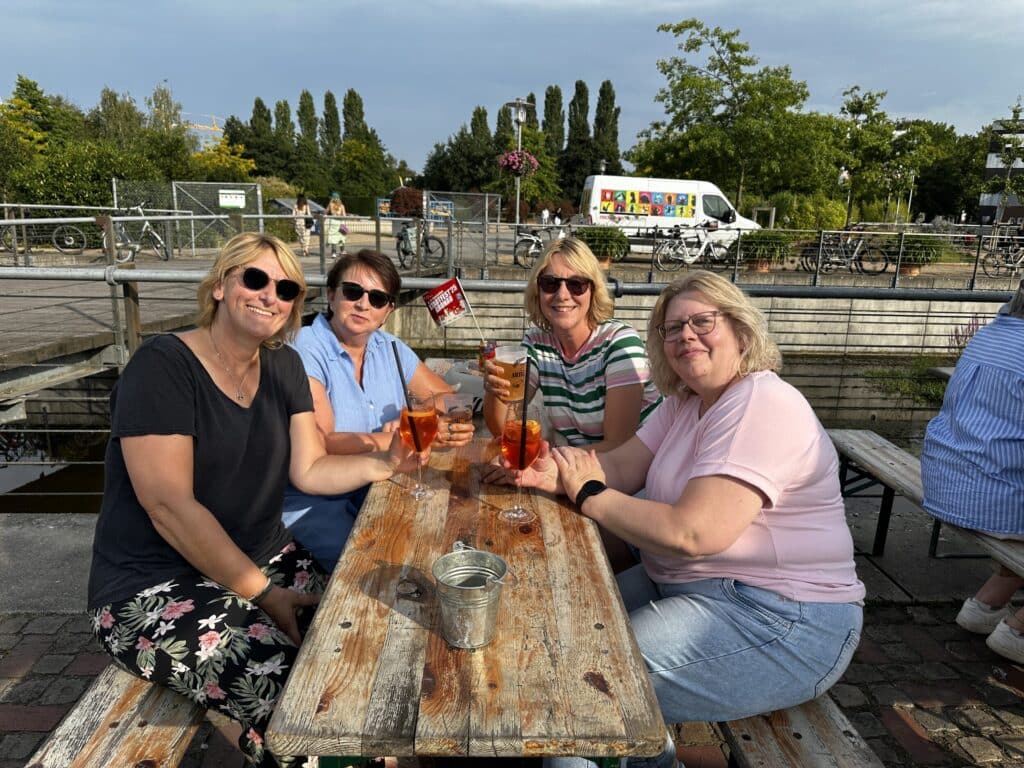 Op de foto zitten vier personen aan een biertenttafel en drinken ze elk een Aperol. Ze glimlachen naar de camera.