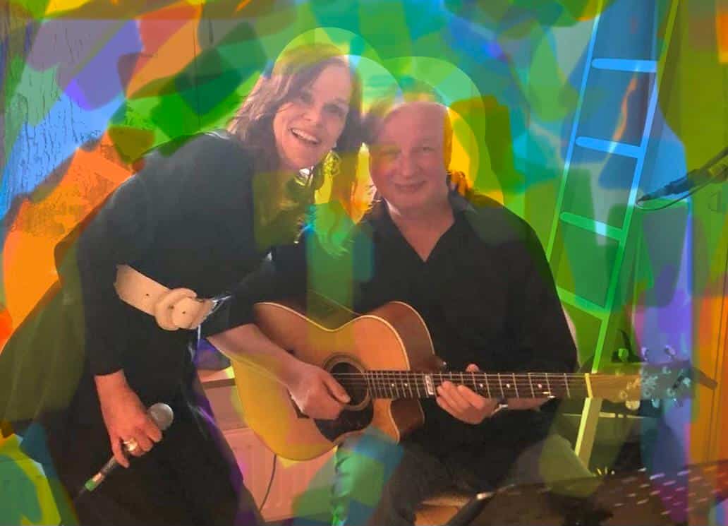 Zwei Menschen stehen eng beieinander in einem bunt beleuchteten Raum. Die Person links hält ein Mikrofon in der Hand und lächelt in die Kamera. Die Person rechts sitzt mit einer Akustikgitarre auf dem Schoß. Über dem ganzen Bild liegen starke farbige Lichtreflexe in Grün, Blau, Gelb und Orange, die eine kreative und lebhafte Konzertstimmung erzeugen. Im Hintergrund ist eine Leiter zu sehen.
