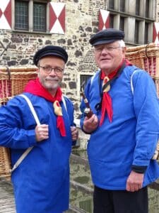 Op de foto zie je twee mannen die verkleed zijn als Kiepenkerle. Ze dragen allebei een blauwe shirt met lange mouwen en een rode sjaal om hun nek. Ze hebben allebei een zwarte pet op.
