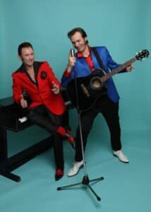 Op de foto zijn de twee artiesten van de Magic Boogie Show te zien. De ene draagt een blauw colbert en houdt een gitaar vast. De andere draagt een rood colbert. Beiden glimlachen naar de camera.
