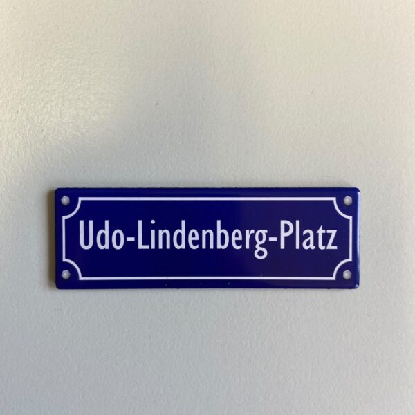 Ein blaues Straßenschild mit weißer Schrift und weißer Umrandung an einer hellen Wand. Darauf steht „Udo-Lindenberg-Platz".