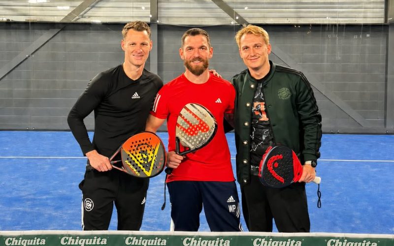 In the picture, you can see three people in athletic clothing standing on a paddle tennis court, each holding a paddle.