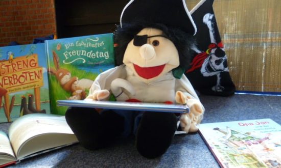 Auf em Bild zu sehen ist das Kuscheltier "Büchereipirat" Pit. Dieser liest aus einem Buch vor.
