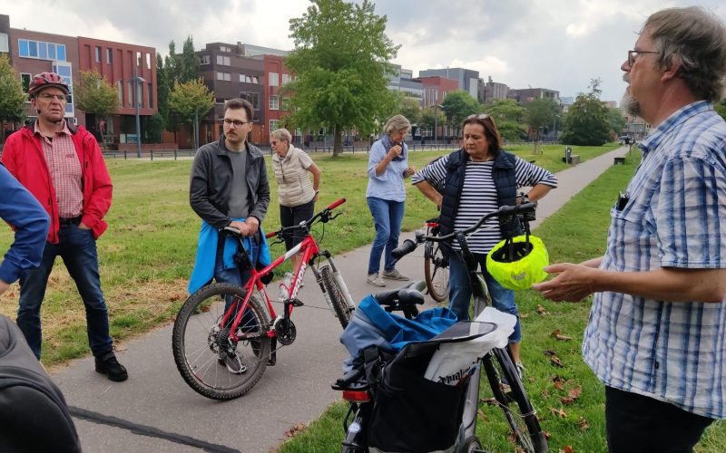 Een groep van zeven volwassenen is gestopt op een fiets- en voetpad. Sommigen staan naast hun fietsen en luisteren naar een man rechts in beeld, die blijkbaar iets uitlegt. Op de achtergrond zijn een groot groen gebied, bomen en moderne gebouwen te zien. De lucht is zwaar bewolkt.