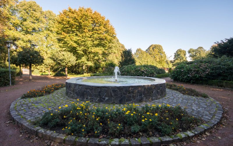 A round stone fountain with water spraying from it stands in the center of a circular flower bed filled with yellow flowers, surrounded by trees and bushes in Eper Park on a clear, sunny day.
