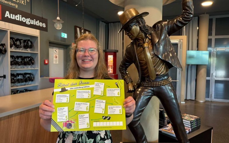 Op de foto is Anne Holters te zien, medewerkster van Stadtmarketing Gronau. Ze staat voor een klein standbeeld van Udo Lindenberg in het museum en houdt de Udo Lindenberg-puzzel in haar hand.
