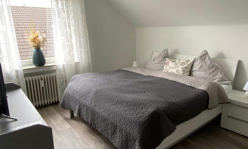Ein helles Schlafzimmer. An der hinteren Wand steht ein Bett, und im Vordergrund ist eine graue Kommode zu sehen.