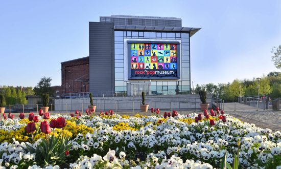 Auf dem Bild zu sehen ist das rock'n'popmuseum aus einer Entfernung von 200 Metern. Vorne im Bild ist ein Blumenbeet mit vielen bunten blühenden Blumen.
